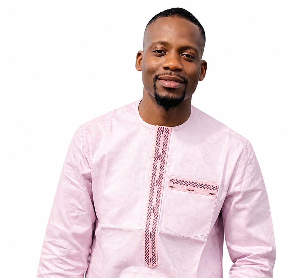 A smiling man wearing a light pink embroidered tunic against a plain white background.