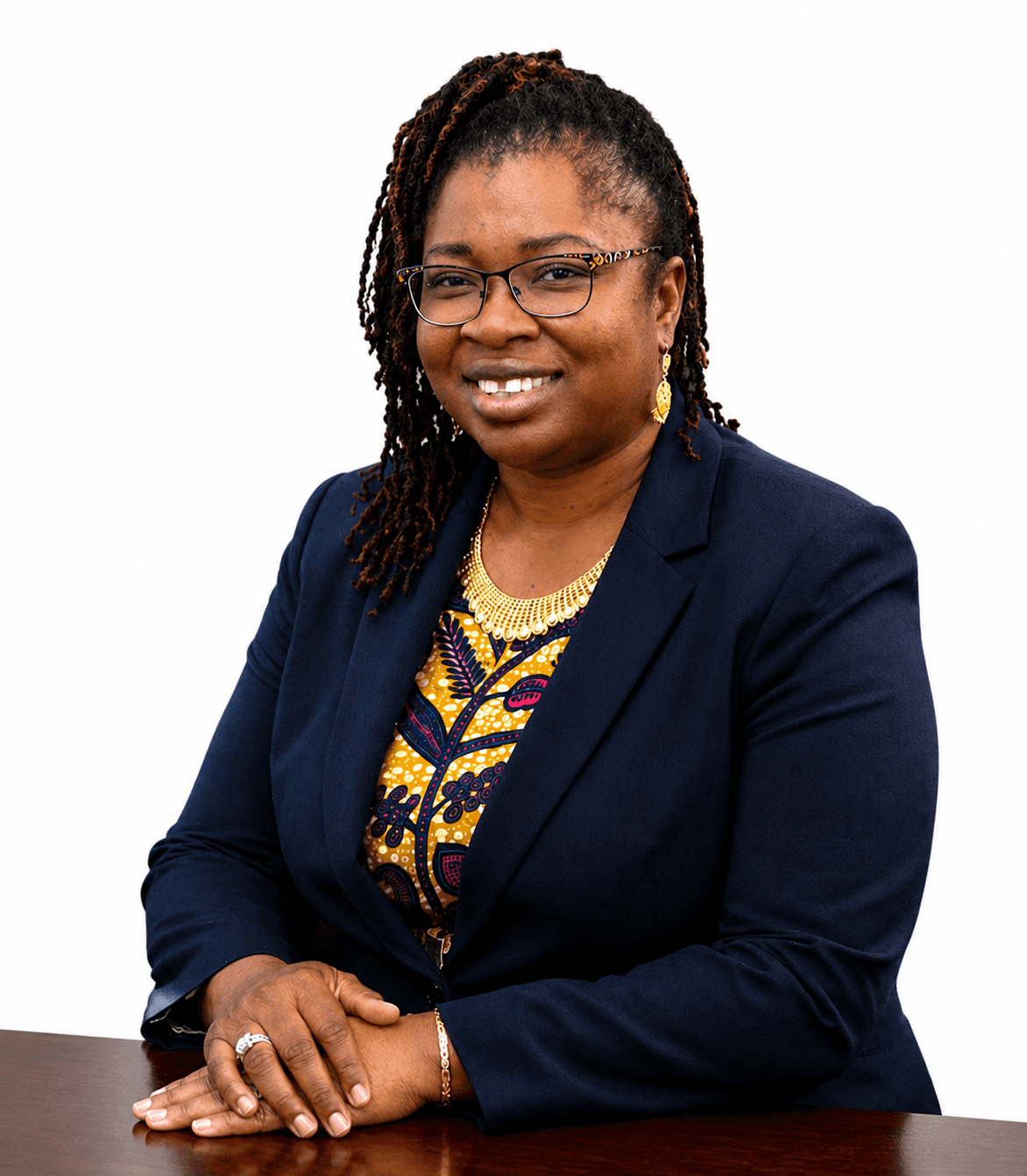 Smiling Black woman with braided hair and glasses wearing a navy blazer at a desk.