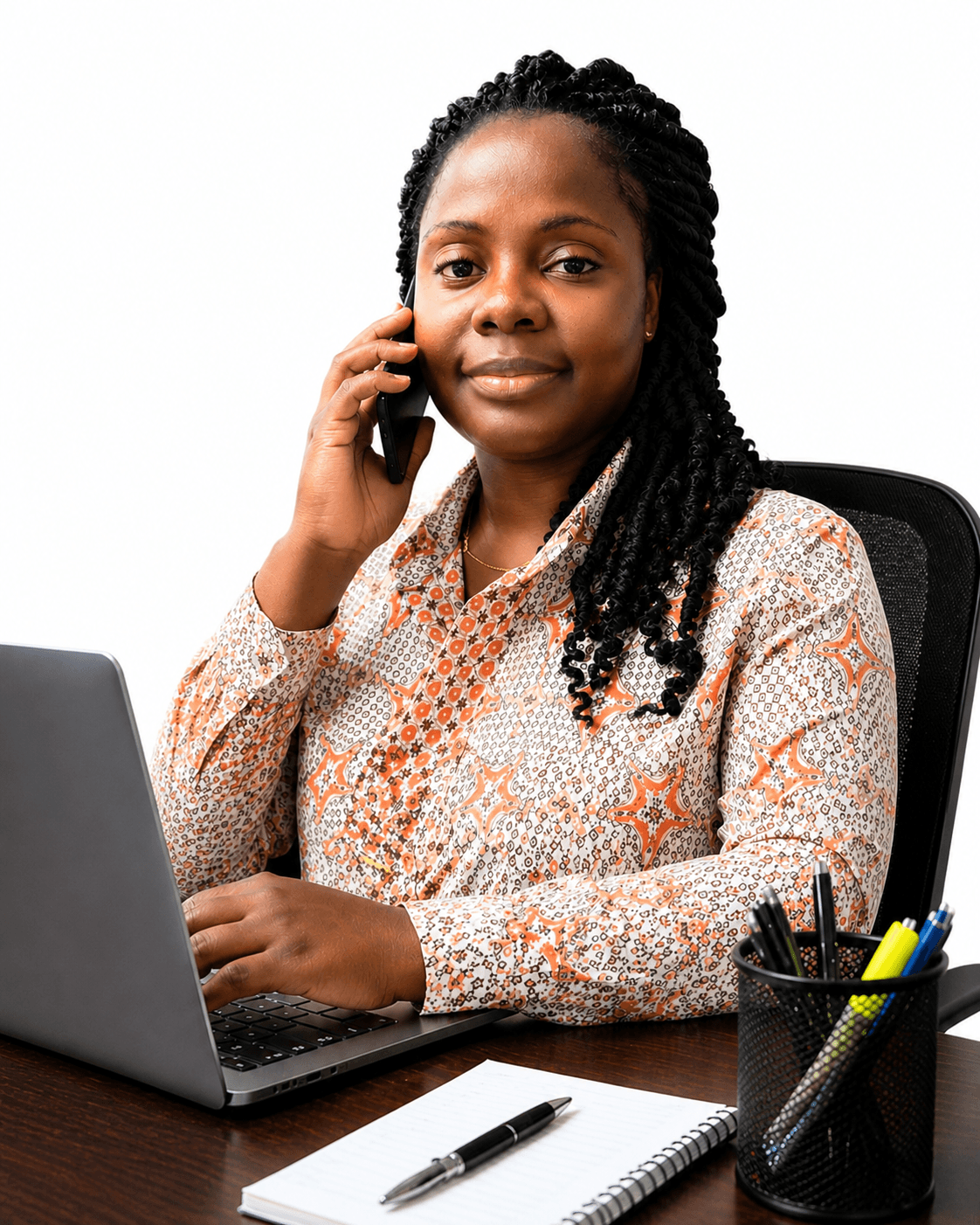 Black woman with braids talking on a phone while using a laptop at a desk.